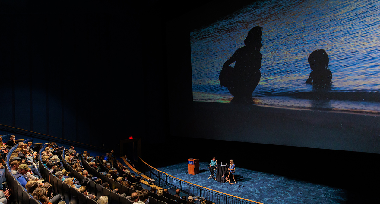 Audience view of the Simons Theater showing a giant screen and people seated in the movie theater seats. Two people are sitting on Q&A seats in front of the screen.